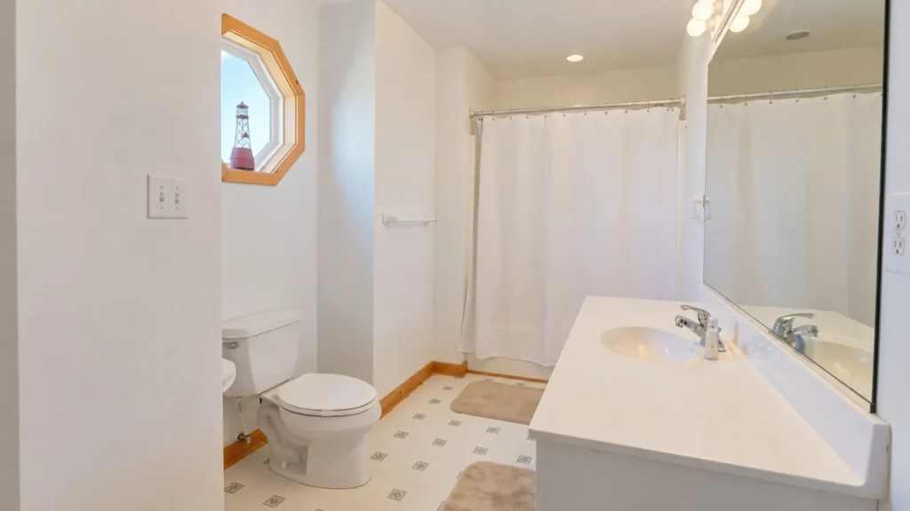 Bright white bathroom with toilet and double-sink vanity, large mirror, white shower curtain, and an octagonal wooden-framed window showing a lighthouse outside.