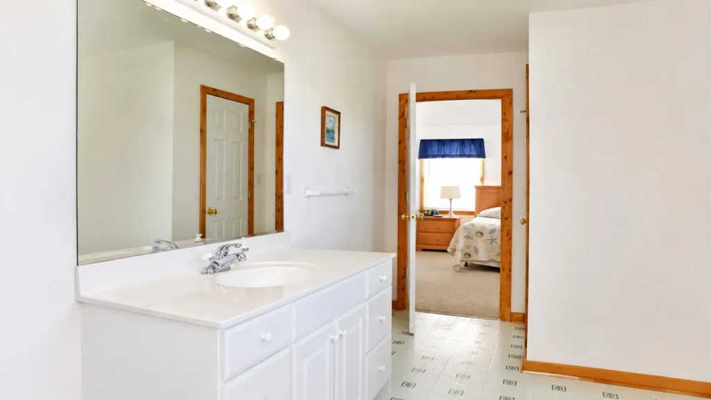 White bathroom vanity with double sink and large mirror, doorway to a bedroom with wooden trim visible in the hall