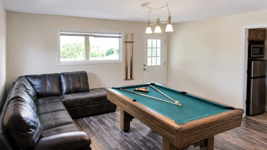 Living room with a pool table and teal felt, a black leather sectional, wall cues, and a door to the outside visible in the background.
