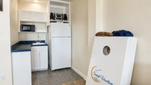 Compact kitchen/utility area with white cabinets, fridge, microwave, sink, and a large laundry machine leaning against the wall in a vacation rental.