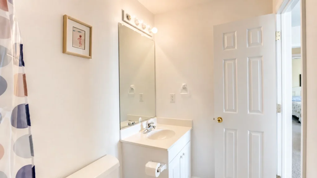 White bathroom with a single-sink vanity, framed art, and a large mirror under a bright light bar, door open to a bedroom beyond the room.