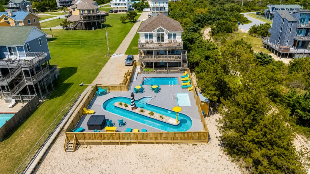 Aerial view of a coastal resort with a curving blue pool, yellow and teal chairs, and a lighthouse-style centerpiece on a sunny deck.
