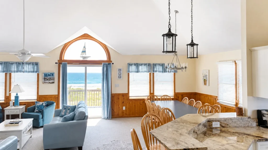 Coastal open-plan living and dining area with blue seating and an ocean view through a large arched window with blue curtains and pendant lights above the table.