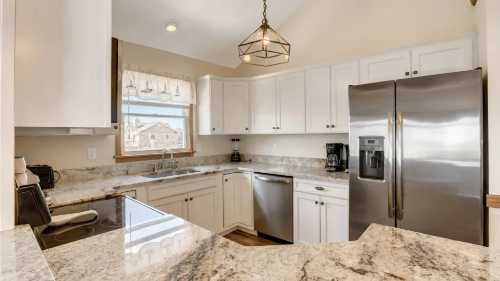 Bright U-shaped kitchen with white cabinets, granite countertops, and stainless steel fridge and dishwasher under a geometric pendant light.