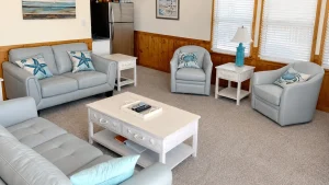Cozy beach-style living room with light blue leather sofa and chairs, white distressed coffee table, and crab/starfish cushions by wooden wainscoting.