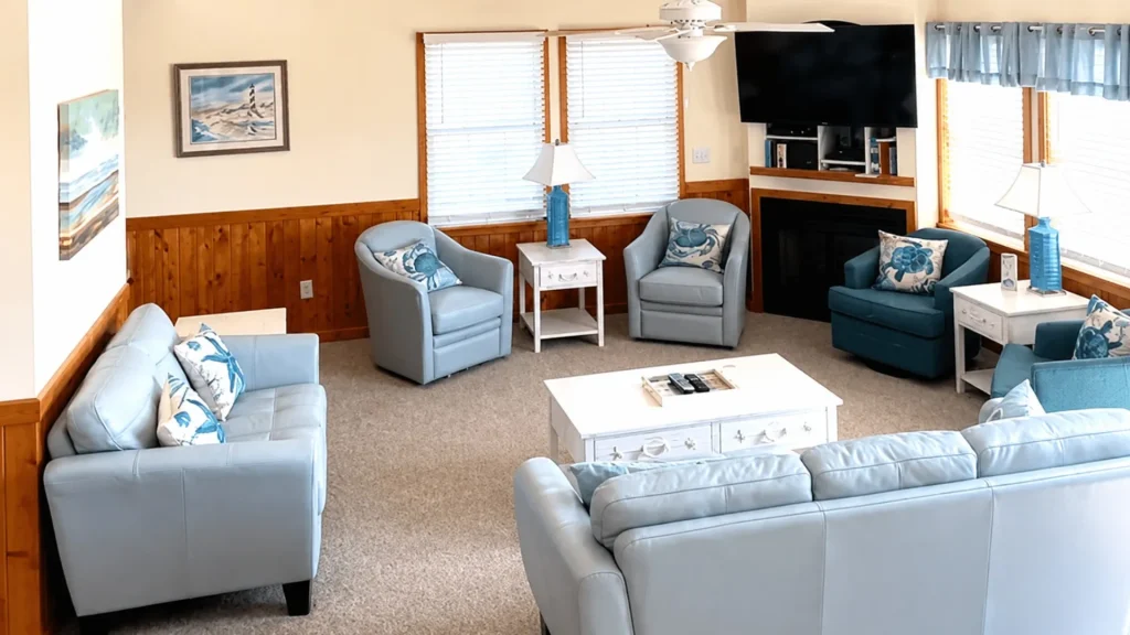 Cozy coastal living room with light-blue seating, white coffee table, wood paneling, a fireplace, and a wall-mounted TV above.