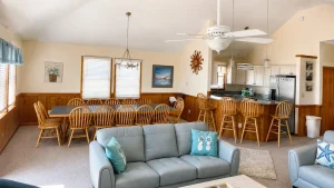 Open-plan family room with a long wooden dining table and many chairs, beige walls, and a white ceiling fan overlooking a kitchen area.