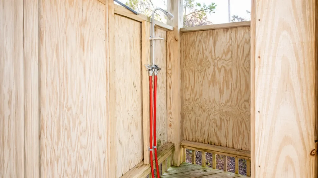 Outdoor wooden shower enclosure with a chrome showerhead and red water pipes connected to a shared valve system inside a partially framed structure.