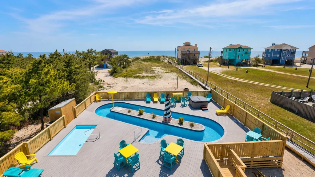 Aerial view of a coastal pool area with turquoise water, colorful chairs, and beach houses beyond the dunes and ocean in the distance.