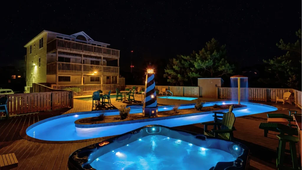Nighttime pool area with glowing blue water, a striped lighthouse centerpiece, and a wooden deck with chairs and a house in the background under a starry sky.