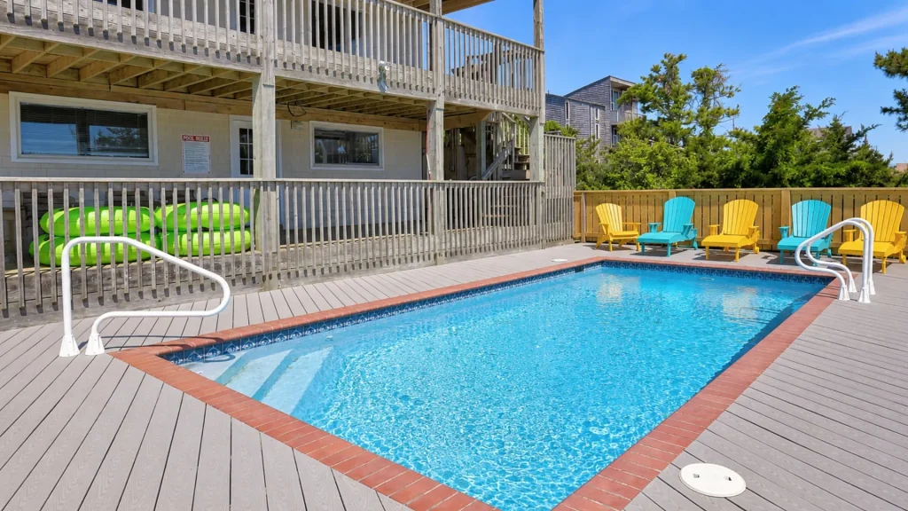 Outdoor pool area with a wooden building and two balconies, lime-green pool tubes stored under the deck, and colorful yellow and turquoise chairs along a fence.