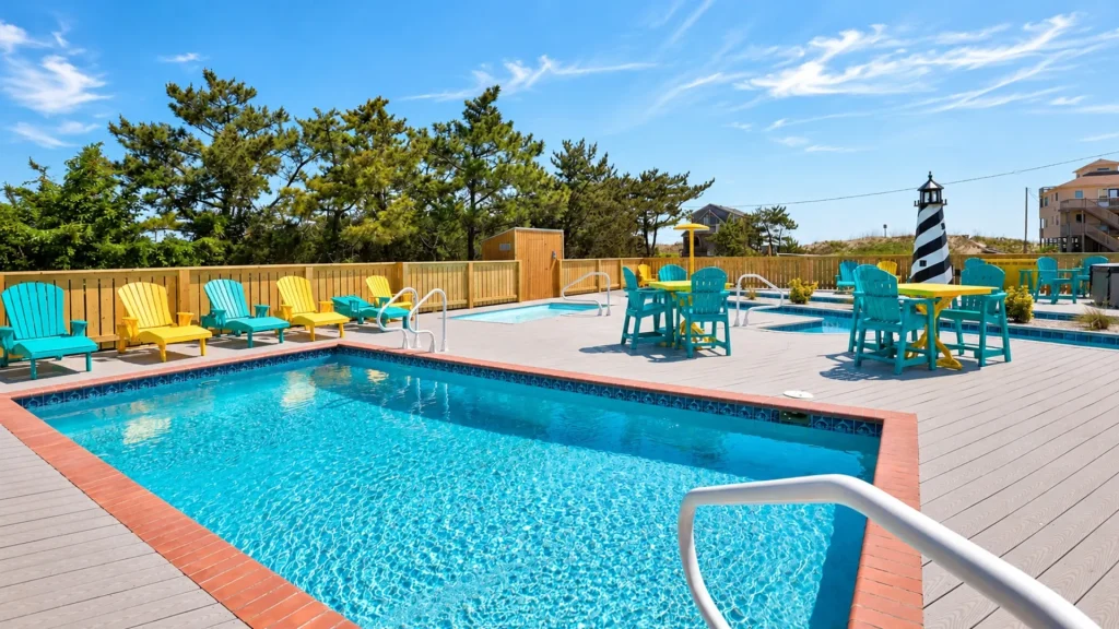Outdoor pool area with blue water, colorful yellow and turquoise chairs, wooden deck, and a striped lighthouse in the background.