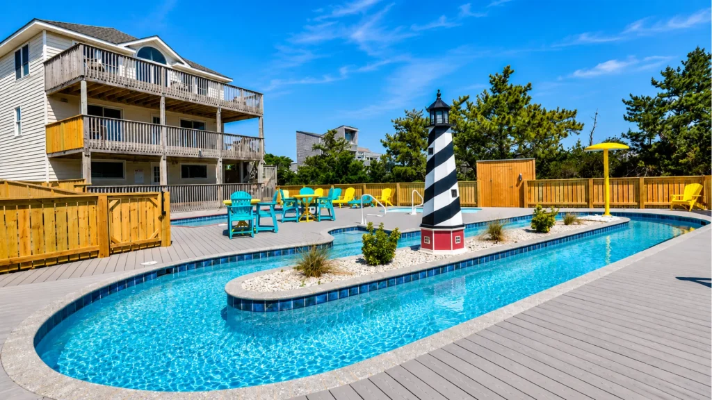 Outdoor pool area with a striped lighthouse centerpiece, blue water, and colorful chairs on a wooden deck near a beige building.