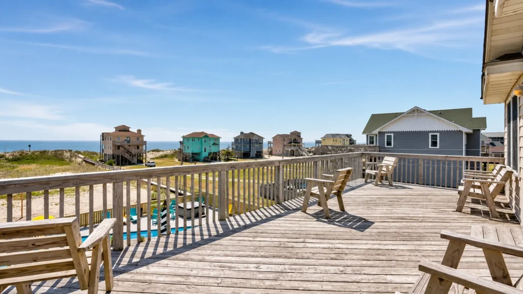 Wooden deck with Adirondack chairs overlooking a row of colorful beach houses and the ocean on a sunny day.