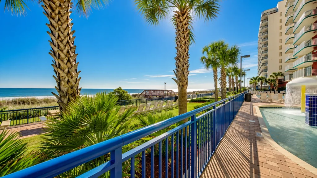 Resort poolside and walkway along a blue railing with palm trees, beach, and ocean in the background.