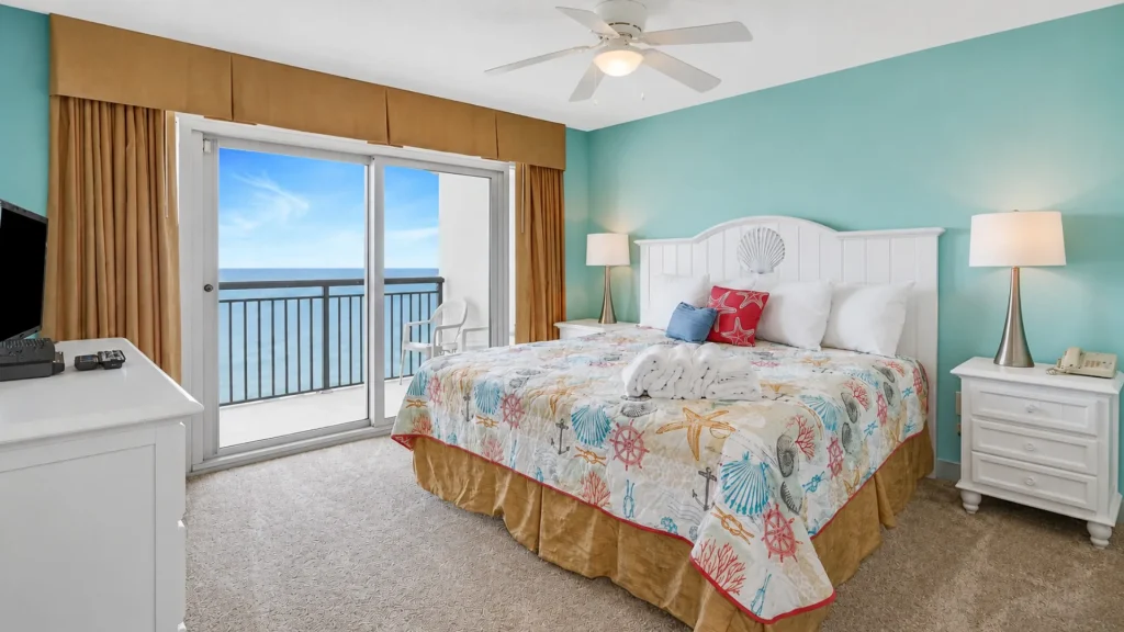 Coastal hotel room with turquoise walls, white headboard bed, seashell quilt, and ocean-view balcony door to the sea beyond.