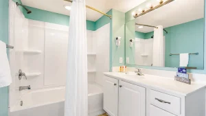 southern-breeze-2-full-bath-2 | Swack Vacations Bright bathroom with teal walls, white tub/shower and shower curtain, and a white double-sink vanity with a large illuminated mirror.