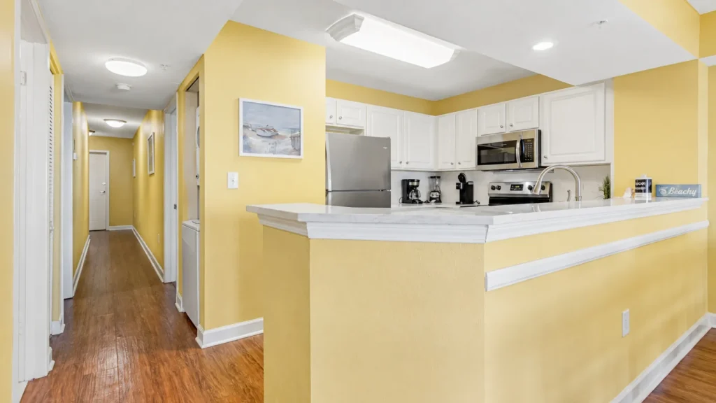 Bright yellow kitchen with white cabinets, stainless steel fridge and microwave, and marble-topped island; hallway to the left.