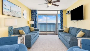southern-breeze-2-living-2 | Swack Vacations Cozy living room with blue couches, yellow walls, and a balcony sea view through sliding doors.