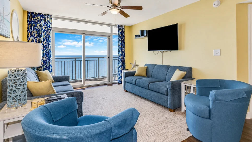 Bright coastal living room with blue sofa set, yellow walls, and a balcony overlooking the ocean with clear skies.