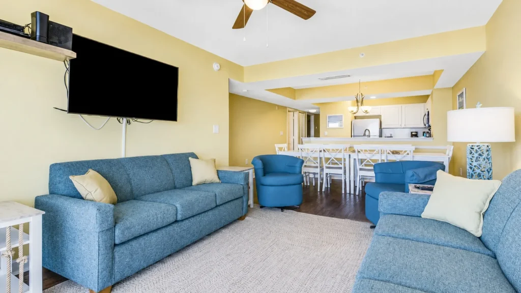 Open-concept living room with blue sofas and chairs, a wall-mounted flat-screen TV, and a dining/kitchen area in the background.