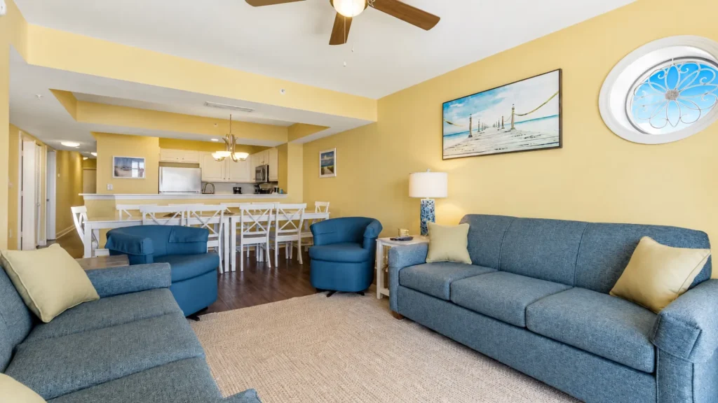 Open-concept living area with yellow walls, blue sofas, and a kitchen bar in the background.