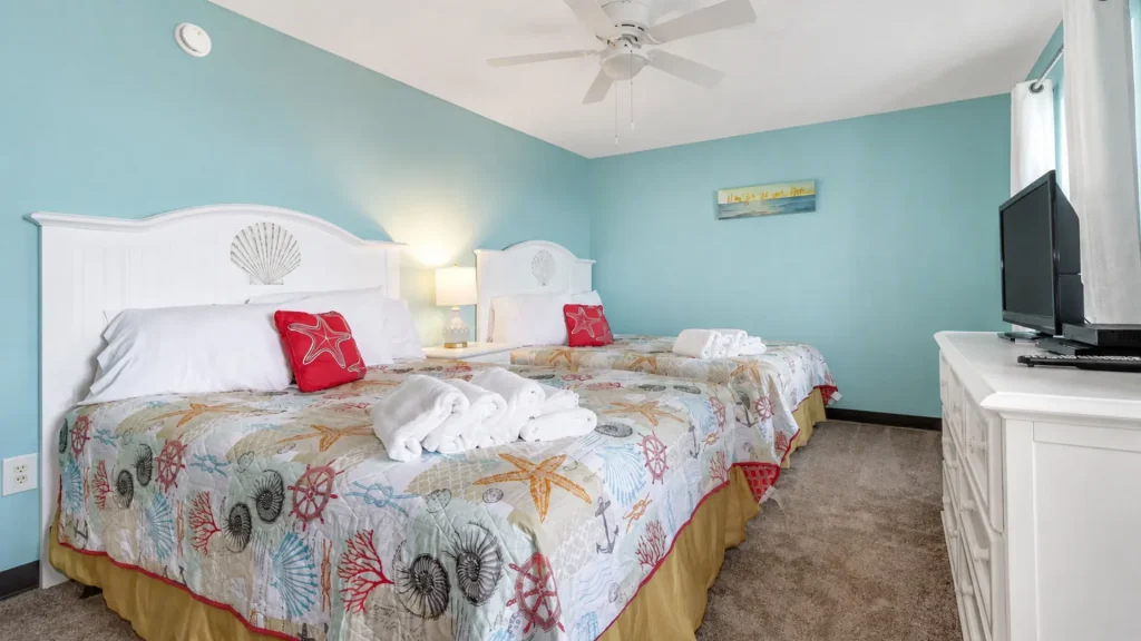 Two white-paneled queen beds with seashell headboards, red starfish pillows, and sea-themed quilts in a bright beach-style hotel room.