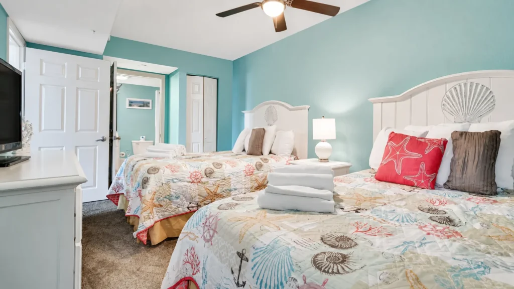 Twin beds with shell-pattern quilts in a bright turquoise beach-themed hotel room with white headboards and pillows.