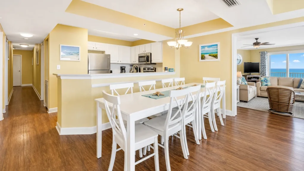 Bright coastal dining area with a long white table and twelve white chairs, yellow walls, and a white kitchen with stainless appliances behind.