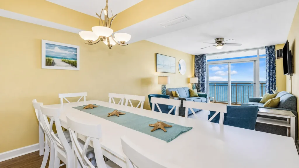 Bright coastal living and dining area with white furniture, yellow walls, and an ocean-view balcony.