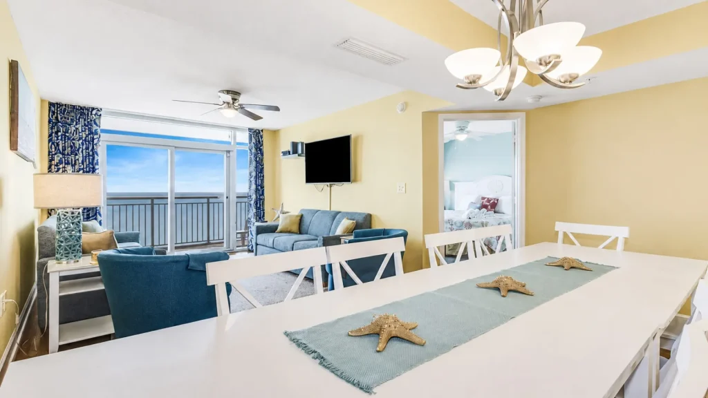 Bright coastal living area with a white dining table and teal table runner decorated with starfish, blue sofas, and a balcony overlooking the ocean.