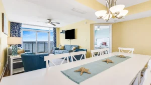 southern-breeze-3-dining-3 | Swack Vacations Bright coastal living area with a white dining table and teal table runner decorated with starfish, blue sofas, and a balcony overlooking the ocean.