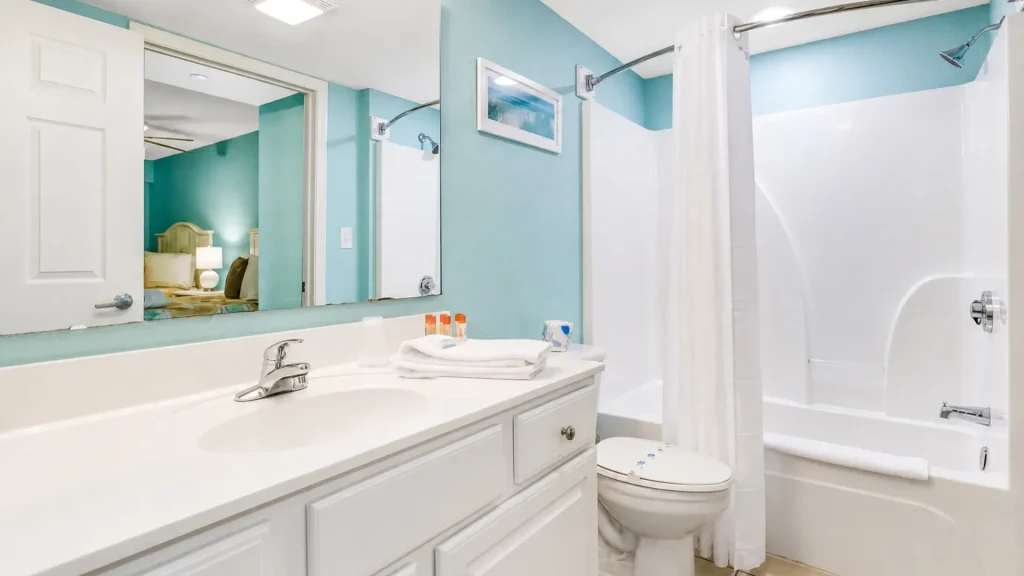 Bright bathroom with teal walls, white vanity and large mirror, folded towels, and toiletries on the counter beside a sink and faucet.