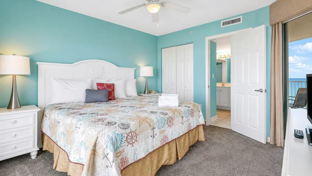 Bright hotel bedroom with teal walls, white headboard, sea-themed quilt, and matching nightstands with lamps.__','King bed in seaside-themed hotel room with ocean-view balcony and turquoise walls.','Coastal hotel room featuring white furniture, beige carpet, and a doorway to the bathroom.','Bed with decorative pillows and a coastal quilt, two lamps, and a balcony door overlooking the sea.','Coastal-themed hotel room with shell-inspired headboard, two lamps, and an ocean-view balcony.