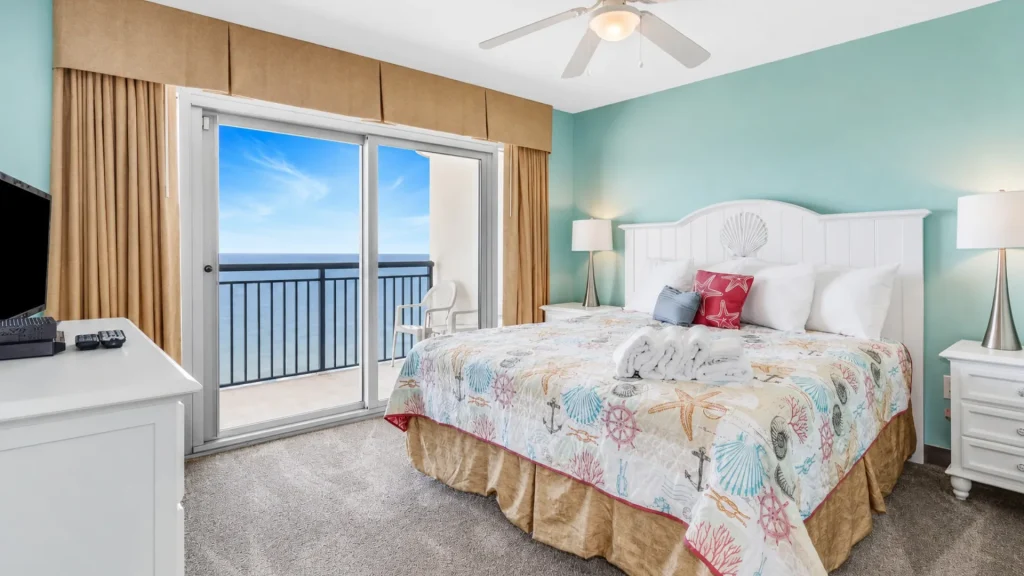 Queen bed with a seashell-pattern quilt in a seaside hotel room, sliding glass doors to a balcony with an ocean view.