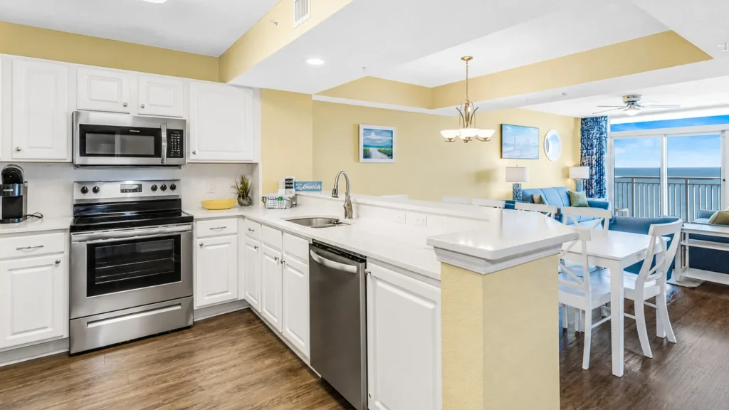 Bright white kitchen with stainless steel stove, microwave, and dishwasher, island sink, abrir to a yellow living area with an ocean-view balcony.