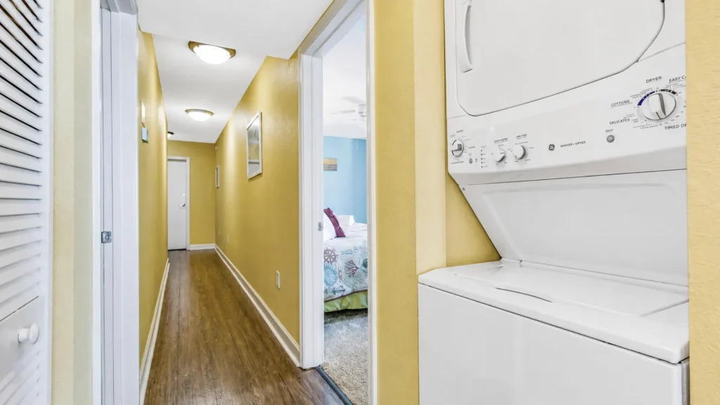 Narrow hallway with yellow walls and wood-like flooring; open doorway reveals a blue bedroom, while a stacked washer and dryer sits on the right wall.