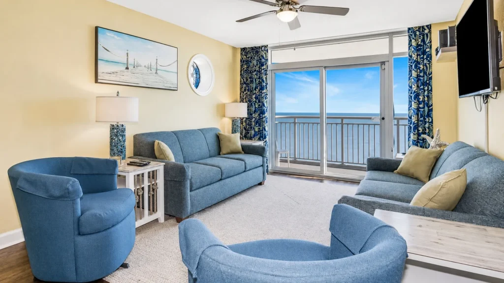 Cozy coastal living room with blue couches, beige walls, and a balcony ocean view through sliding glass doors.