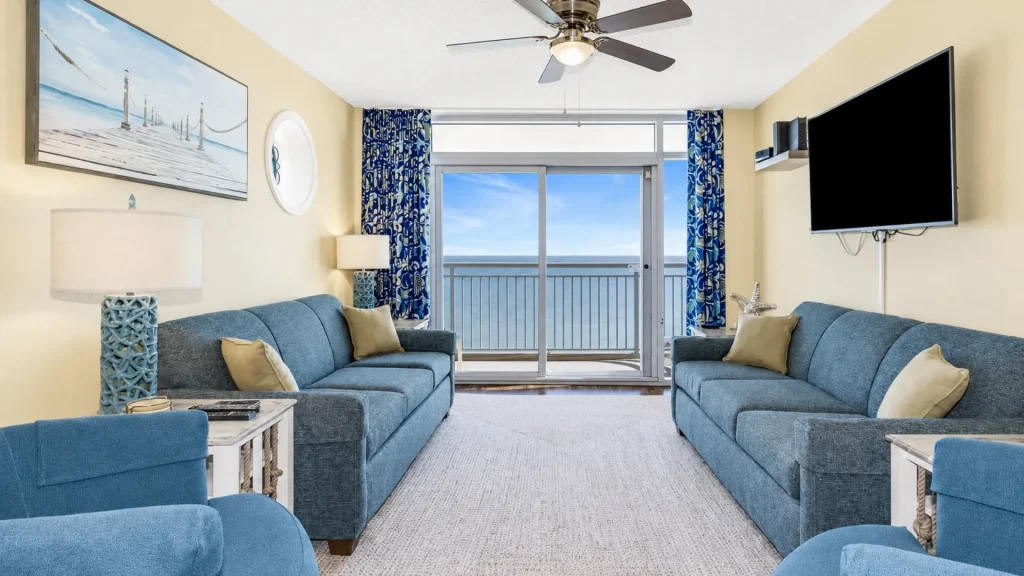 Coastal living room with blue sofas, beige walls, and a sliding door open to a balcony overlooking the ocean.
