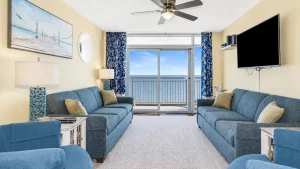 southern-breeze-3-living-2 | Swack Vacations Coastal living room with blue sofas, beige walls, and a sliding door open to a balcony overlooking the ocean.