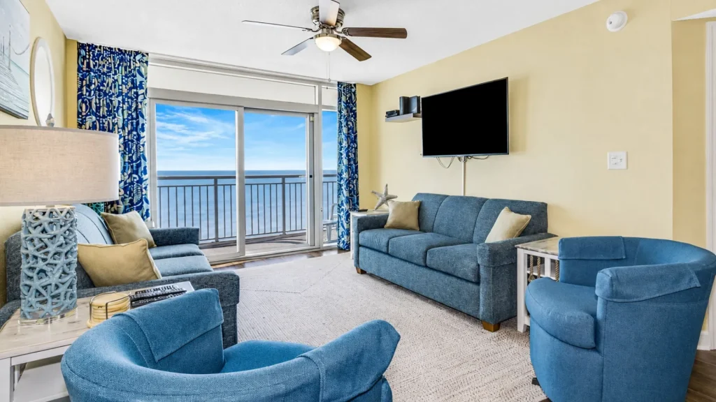Coastal living room with blue upholstered sofa and chairs, and a balcony overlooking the ocean through sliding doors.