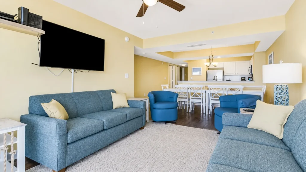 Open living room with blue sofas and a wall-mounted TV; yellow walls and a white dining table in the background.