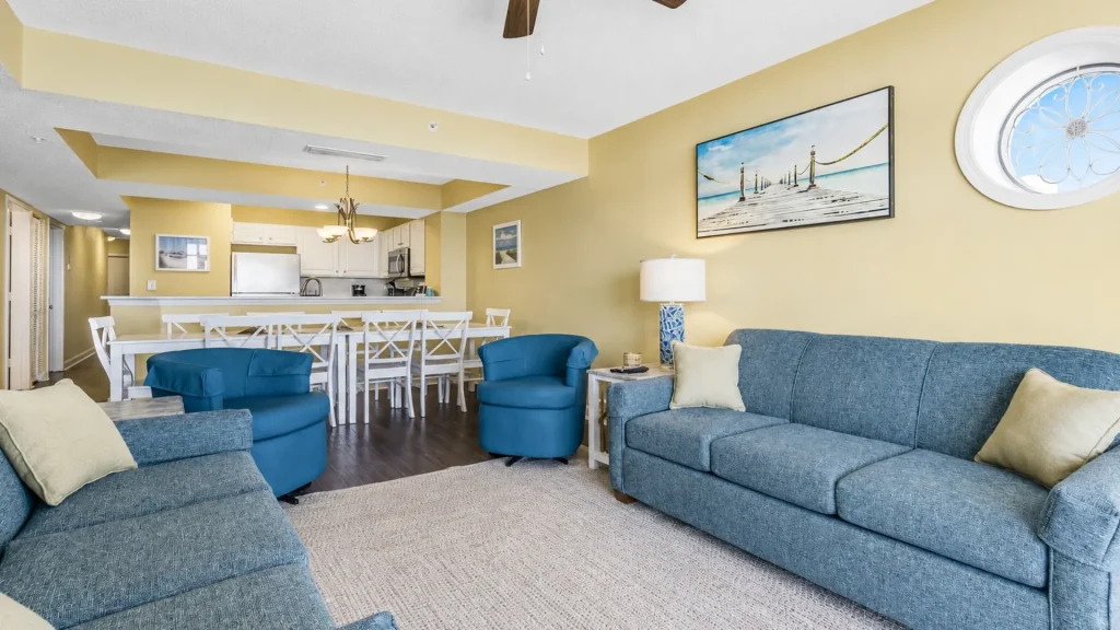 Open-concept living room with blue sofas and chairs, yellow walls, and a white kitchen in the background.