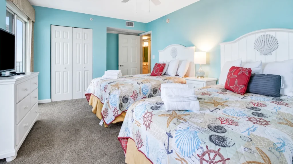 Coastal hotel room with two beds, shell-themed headboards, and turquoise walls; dresser with TV on the left and a nightstand lamp between the beds