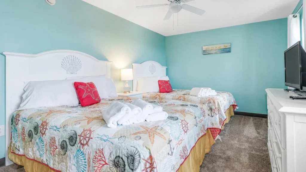 Two white headboard beds with sea-life quilts, red starfish pillows, and folded towels in a bright beach-themed hotel room.