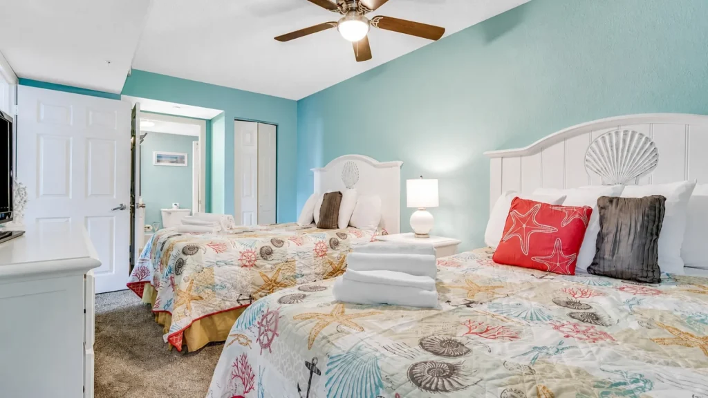 Bright coastal hotel room with two beds, turquoise walls, white headboards with shell detail, and a ceiling fan.