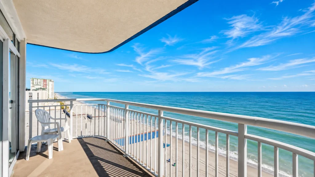 Balcony view overlooking a sandy beach and blue ocean, with a white chair and railing in the foreground.