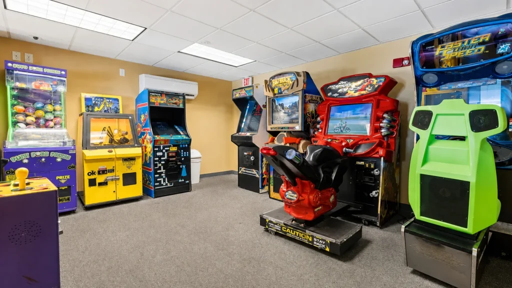 Colorful arcade game room with various upright and pedestal machines along beige walls and carpeted floor.