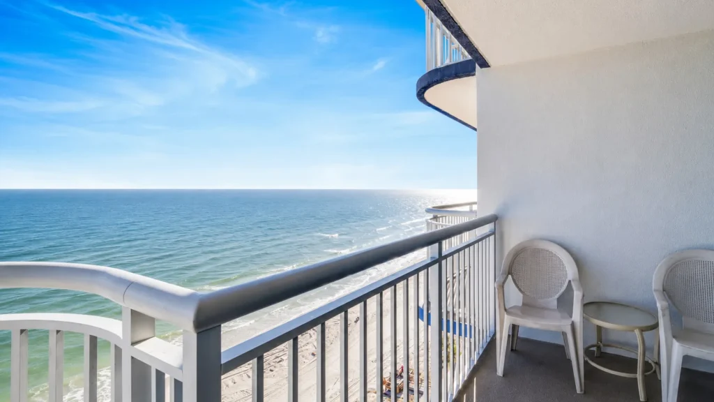 Balcony with two plastic chairs and a small table overlooking blue ocean and sandy beach far below.