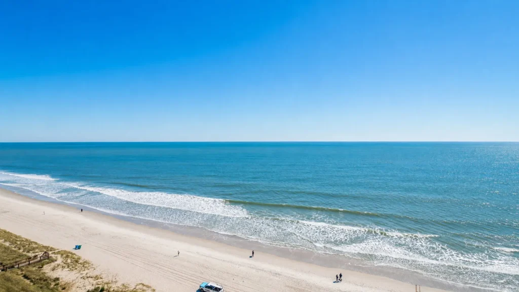 Wide sandy beach meeting calm blue ocean under a clear blue sky at horizon line. A few people and a vehicle are visible along the shore.